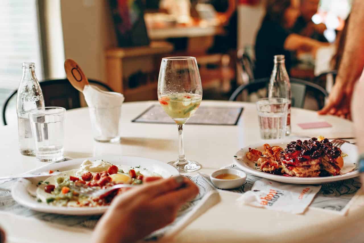 A table set with two plates of food, one containing a colorful dish with vegetables and the other with pancakes topped with berries, a glass of white wine sits between them, accompanied by a water glass and a bottle in the background, creating a cozy dining scene with a blurred background