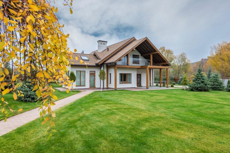 Modern white house with wooden accents and steep roof set on manicured lawn with yellow autumn leaves and evergreens