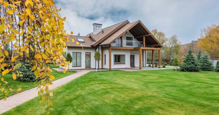 Modern white house with wooden accents and steep roof set on manicured lawn with yellow autumn leaves and evergreens