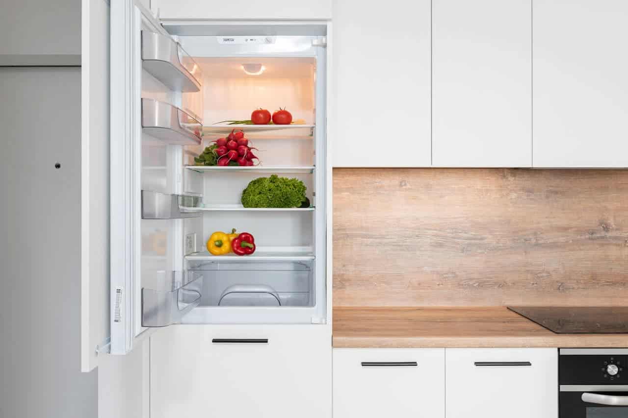 Open refrigerator stocked with fresh vegetables including tomatoes, radishes, bell peppers, and lettuce in a modern white kitchen