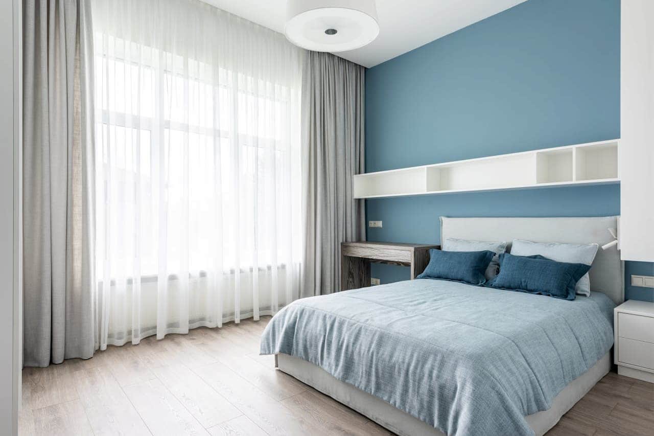 Modern bedroom with light blue accent wall, white bed frame with matching blue bedding, floating shelves, large windows with sheer and opaque curtains, circular ceiling light, and light wood flooring