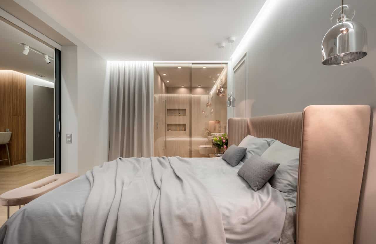 Modern bedroom with pink upholstered headboard, light gray bedding, and glass-enclosed bathroom. Features pendant lighting, recessed ceiling lights, white curtains, and wood accents. Clean minimalist design with soft neutral color palette