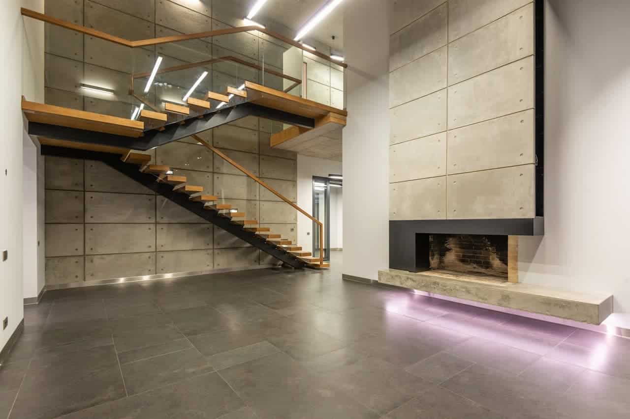 Modern industrial living space with floating wooden staircase, concrete walls, glass railings, dark tile flooring, LED lighting and built-in fireplace