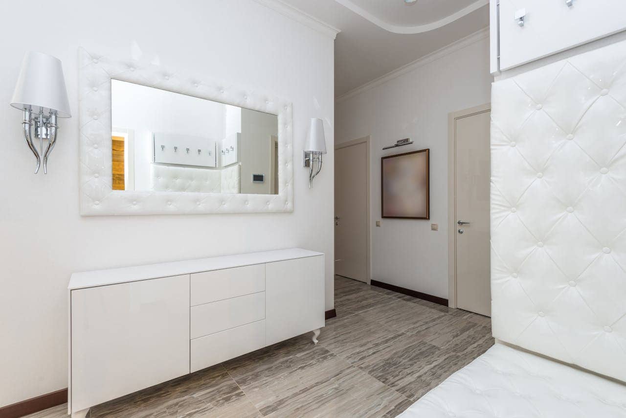 A clean, modern hallway with a white tufted mirror, wall-mounted light fixture, a white dresser with drawers, beige tiled floor, a white door with a simple frame, and an abstract art piece on the wall