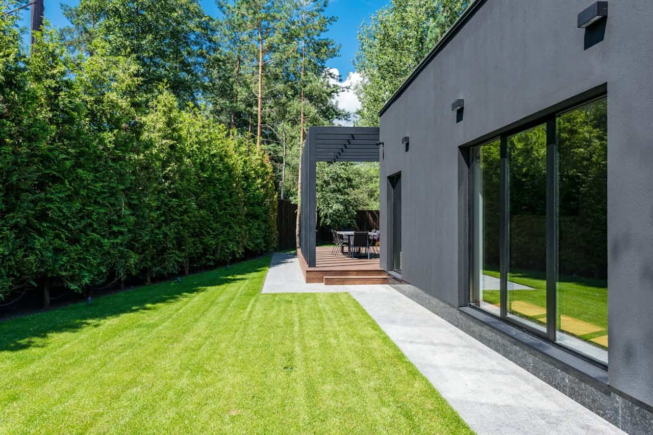 A modern house with a sleek dark exterior, featuring large windows and a wooden deck with outdoor seating, surrounded by a well-manicured lawn and tall green hedges, creating a serene and spacious backyard setting, clear blue skies and lush greenery in the background
