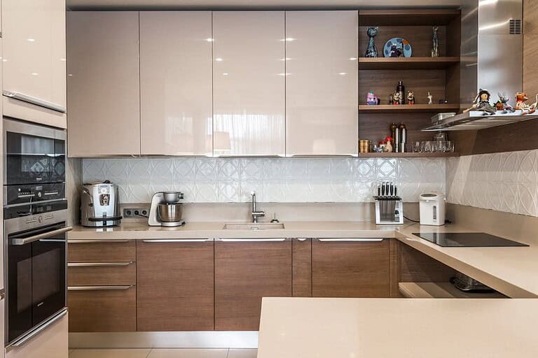 Modern L-shaped kitchen with glossy white upper cabinets, wooden lower cabinets, beige countertops, built-in appliances, and decorative shelving