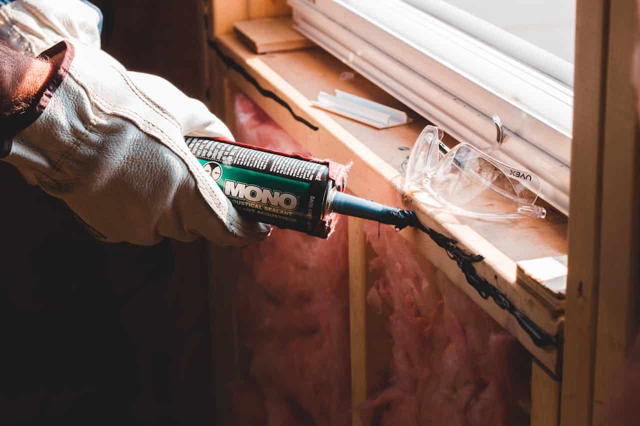 A person wearing white work gloves is applying black sealant from a MONO industrial sealant tube onto a wooden window frame, with safety glasses placed nearby, and insulation material visible in the background