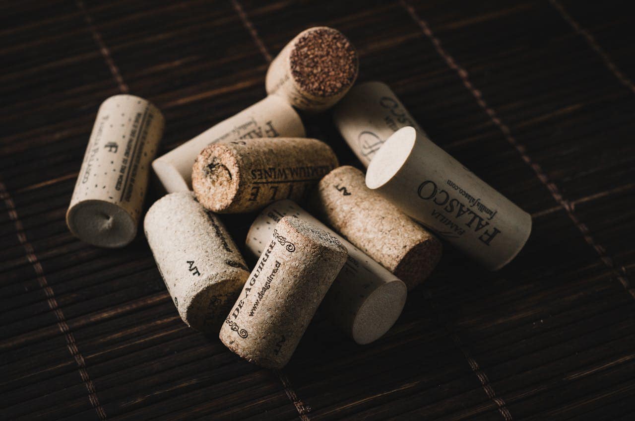 A collection of wine corks scattered on a dark woven surface, displaying printed labels and logos on the corks, some corks with visible markings and others plain, creating a close-up view of wine corks as an aesthetic item