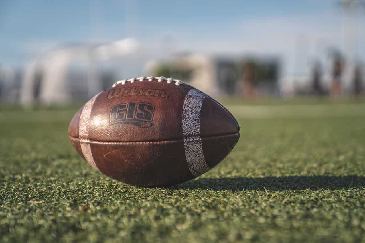 A close-up shot of a Wilson GIS branded football placed on a green grass field, blurred background with indistinct figures, bright lighting illuminating the ball, the ball is in focus with visible texture and stitching