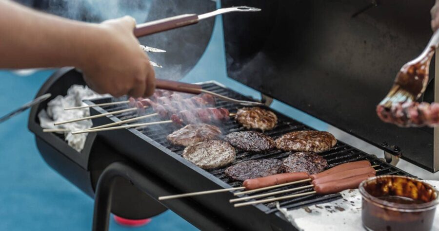 The image shows a barbecue grill with sizzling hamburgers, hotdogs on skewers, and skewers being brushed with sauce, smoke rising from the grill, a person using tongs to flip the burgers, outdoor cooking scene with grilling food, summer barbecue gathering