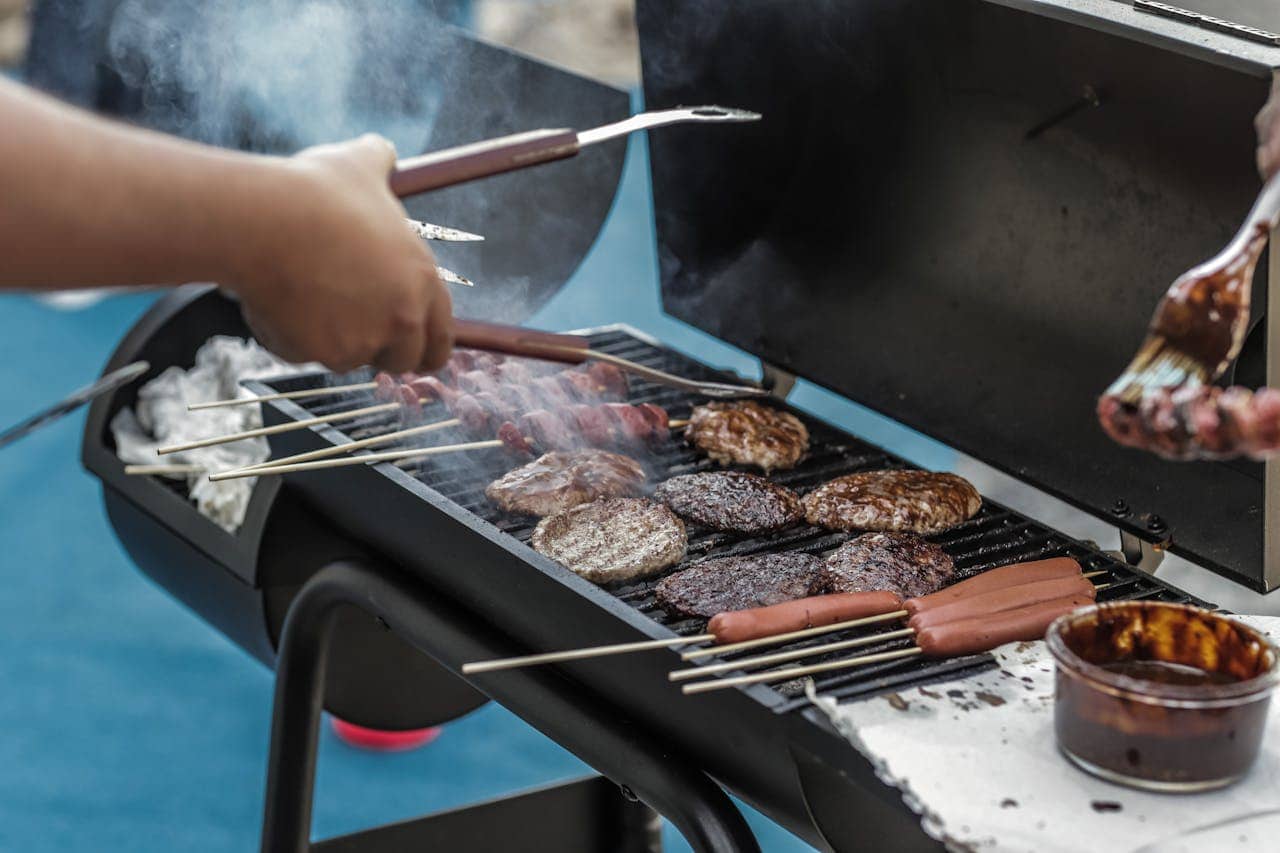 The image shows a barbecue grill with sizzling hamburgers, hotdogs on skewers, and skewers being brushed with sauce, smoke rising from the grill, a person using tongs to flip the burgers, outdoor cooking scene with grilling food, summer barbecue gathering