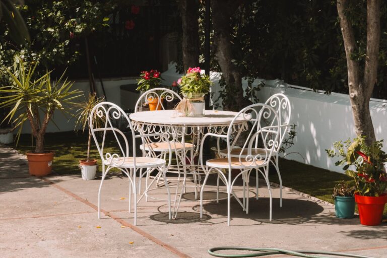 A white metal garden table with four matching chairs, potted plants on the table and around it, green background, a sunny outdoor setting, hose visible on the ground, inviting and peaceful atmosphere with bright natural light