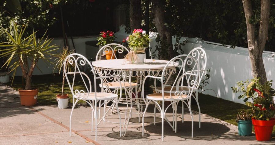 A white metal garden table with four matching chairs, potted plants on the table and around it, green background, a sunny outdoor setting, hose visible on the ground, inviting and peaceful atmosphere with bright natural light