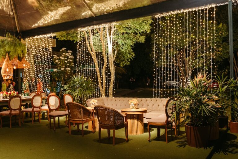 Outdoor dining area with tufted sofa, wicker chairs, string lights hanging as curtains, and tropical plants surrounding the space