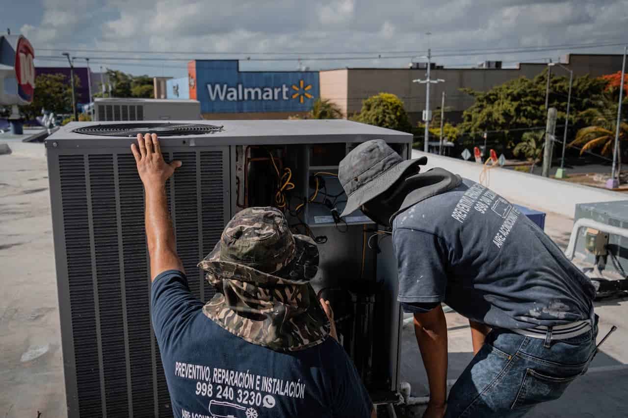 Two HVAC technicians repairing air conditioning unit on rooftop with Walmart store visible in background under cloudy sky