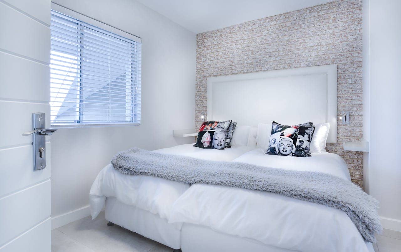 Compact white bedroom with brick accent wall, large white headboard, crisp white bedding, gray throw blanket, and decorative Marilyn Monroe pillows. Features window with horizontal blinds and modern door handle
