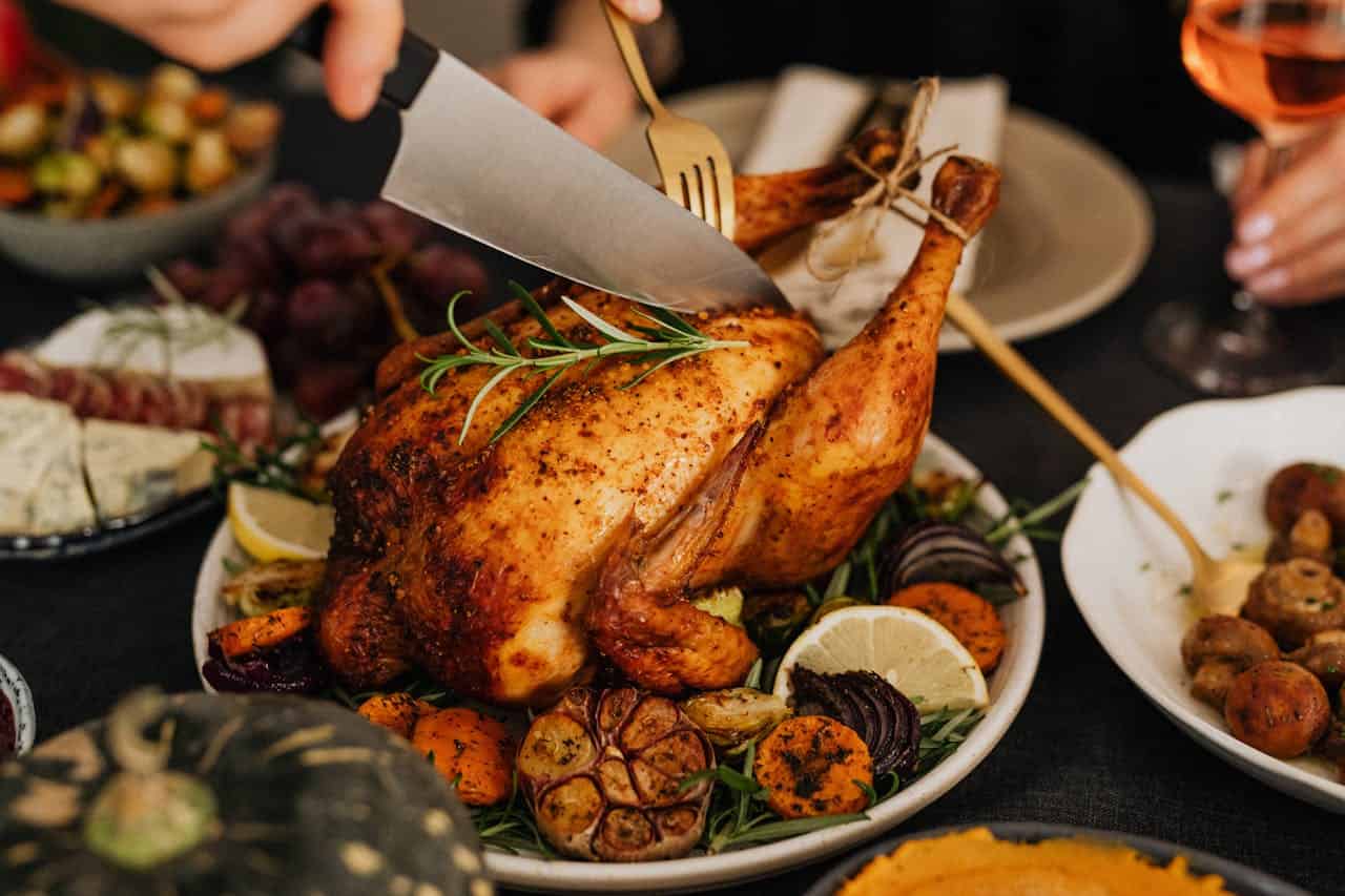 Roasted whole chicken being carved with knife and fork, served with roasted vegetables and garnished with herbs on white plate