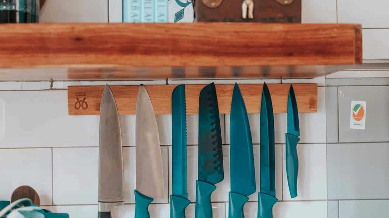 A wooden knife rack with various kitchen knives of different sizes and types hanging, blue-handled knives prominently displayed, a few metallic blades without handles, set against a neutral kitchen background with tiles and a certification sticker visible on the wall