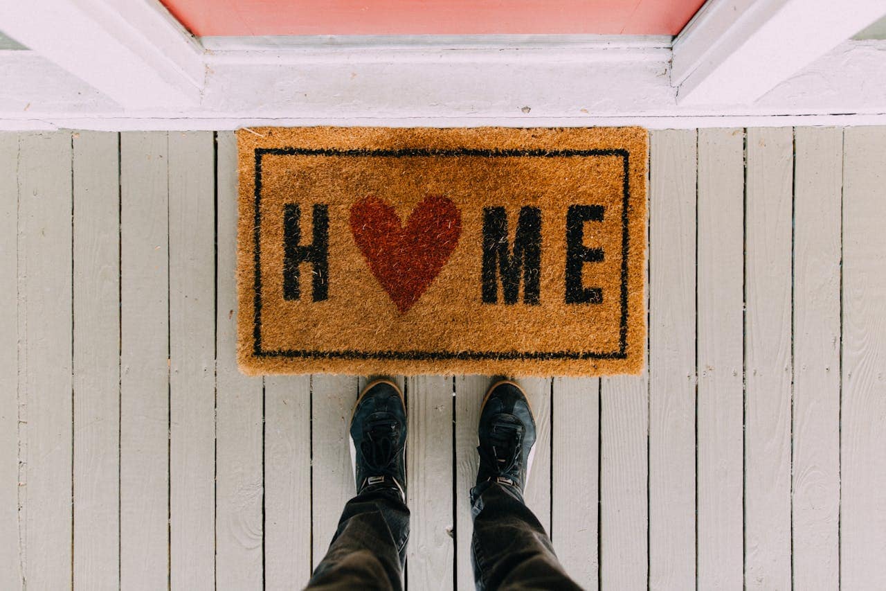 A doormat with the word "HOME" spelled out, a red heart symbol between the letters H and ME, someone standing in front of the door with black sneakers, wooden floorboards visible around the mat
