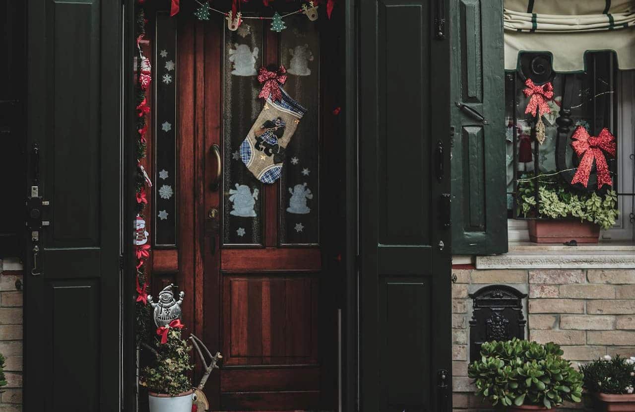 A wooden door with green shutters, decorated with Christmas-themed garlands, a stocking, and snowflake patterns on the glass, red bows on the window, potted plants with greenery, and a festive snowman figurine placed at the base