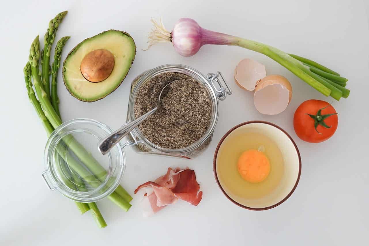 A variety of fresh ingredients on a kitchen countertop, including avocado halves, asparagus, a tomato, green onions, an egg in a bowl, slices of bacon, a jar of black pepper with a spoon, and eggshells, arranged neatly on a light surface ready for cooking or meal prep