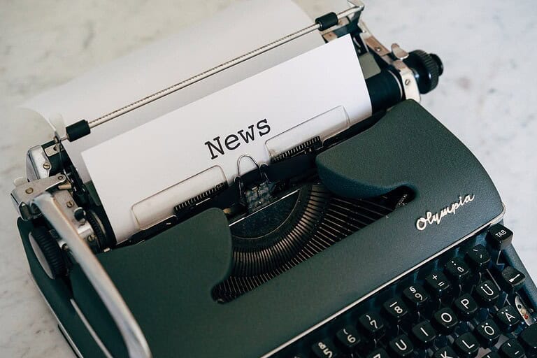 A vintage Olympia typewriter with the word "News" typed on a white sheet of paper, the typewriter keys visible, the machine placed on a light surface