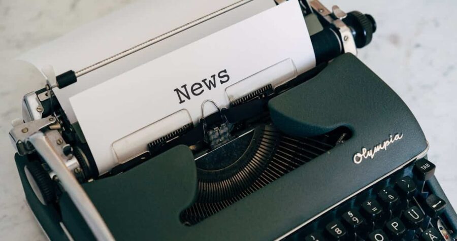 A vintage Olympia typewriter with the word "News" typed on a white sheet of paper, the typewriter keys visible, the machine placed on a light surface