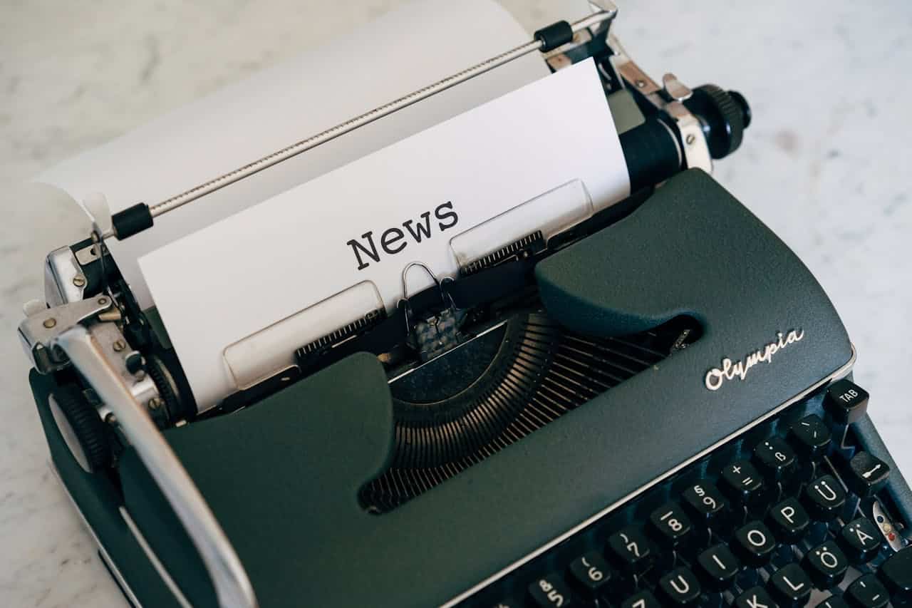 A vintage Olympia typewriter with the word "News" typed on a white sheet of paper, the typewriter keys visible, the machine placed on a light surface