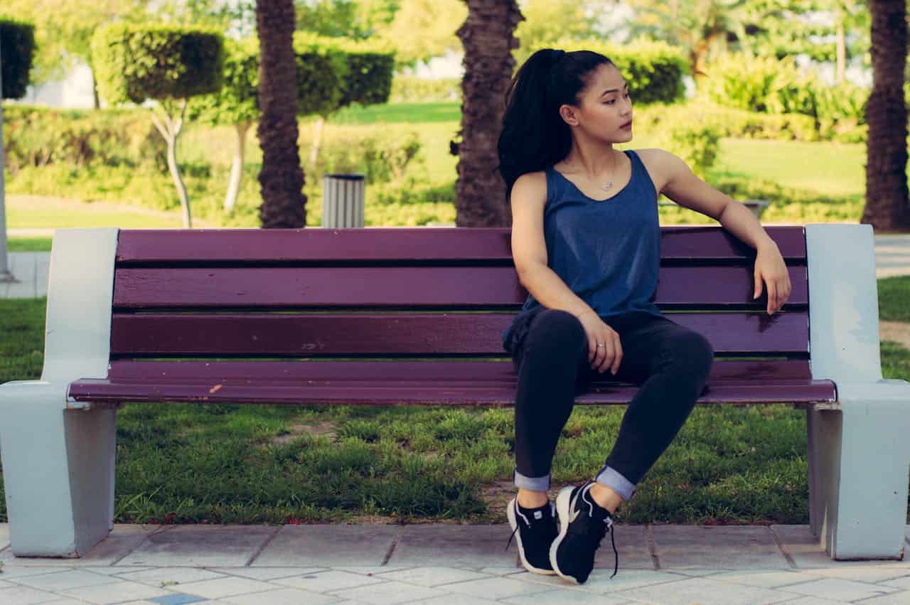 A young woman sitting on a purple park bench in casual attire, with a relaxed posture and ponytail, looking to her right, wearing black pants and sneakers, surrounded by green grass and trees in a well-maintained park, creating a peaceful and serene outdoor scene