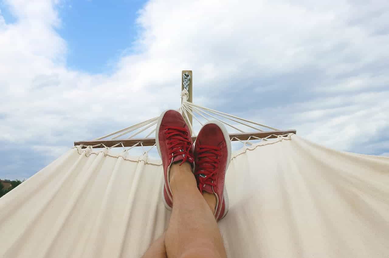 A pair of feet wearing red sneakers resting on a hammock, with the hammock stretched out under a partly cloudy sky, the image captures a moment of relaxation and leisure in an outdoor setting