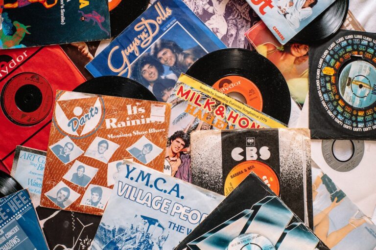 A collection of vinyl records and their colorful album covers scattered on a surface, featuring titles like "Y.M.C.A." by Village People, "It's Raining" by Darts, and "Milk & Honey" with a vintage 45 RPM record, showcasing a nostalgic music collection