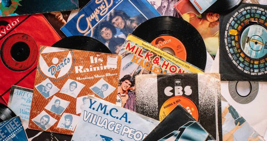 A collection of vinyl records and their colorful album covers scattered on a surface, featuring titles like "Y.M.C.A." by Village People, "It's Raining" by Darts, and "Milk & Honey" with a vintage 45 RPM record, showcasing a nostalgic music collection