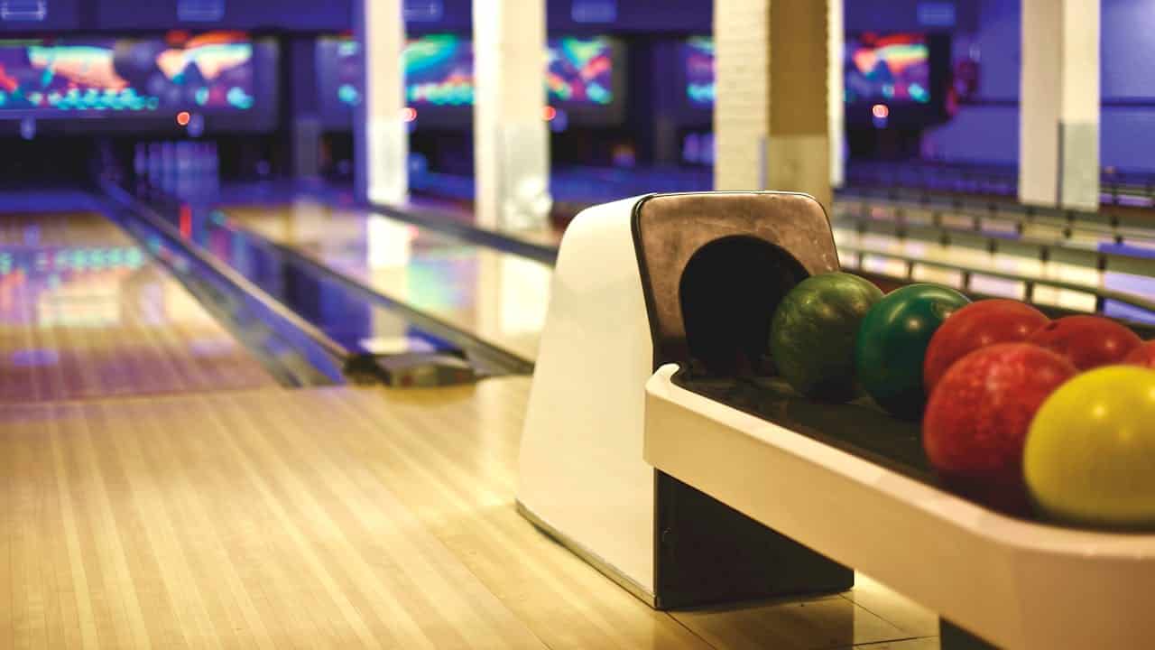 A close-up view of a bowling alley showing a rack of colorful bowling balls, with one yellow ball visible in the foreground, the wooden lanes illuminated by neon lights