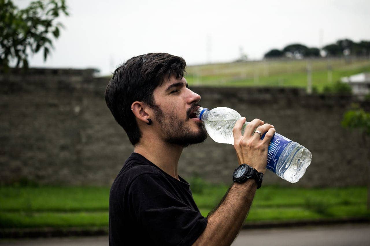 Young man with dark hair and beard drinking from plastic water bottle outdoors, wearing black shirt and digital watch