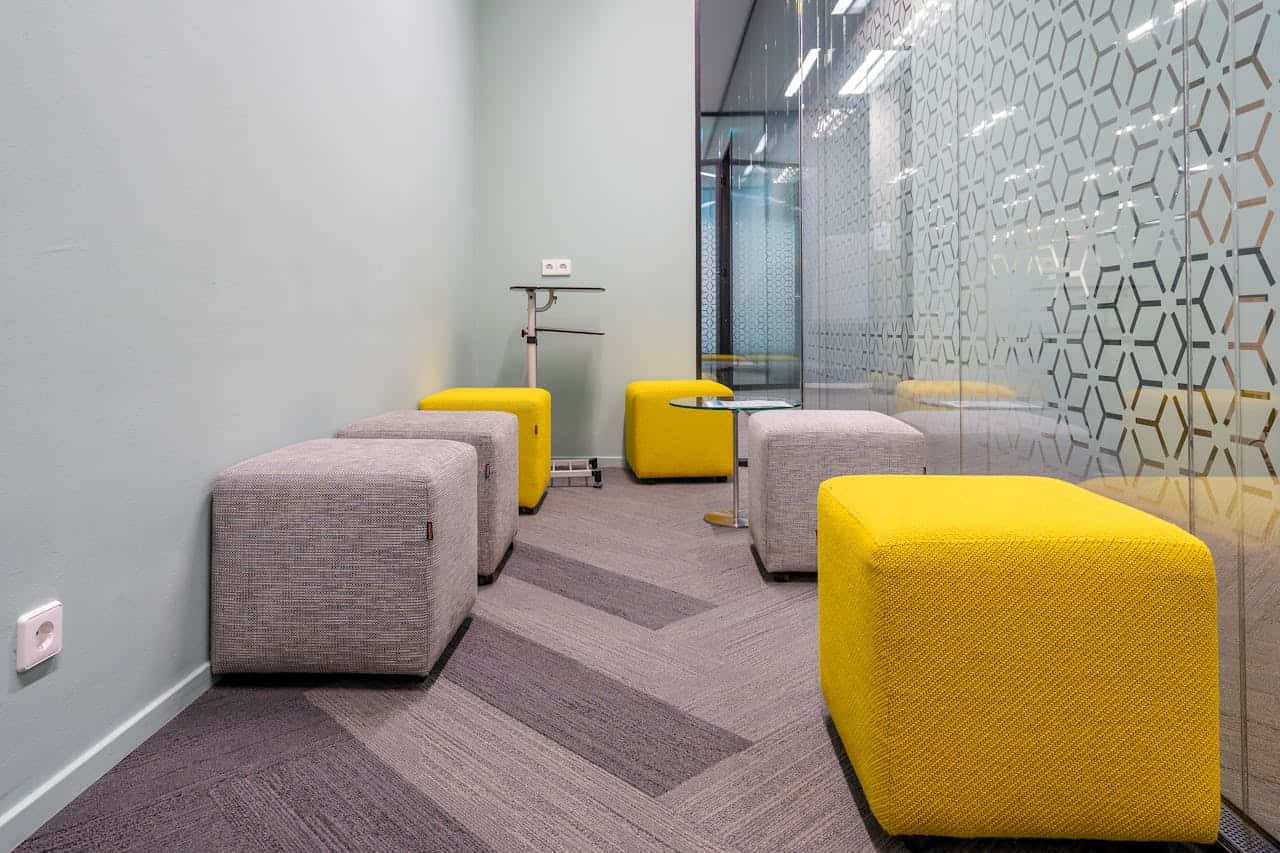 Modern office breakout space with yellow and gray cube ottomans, mint green walls, patterned glass partition, and gray carpet tiles