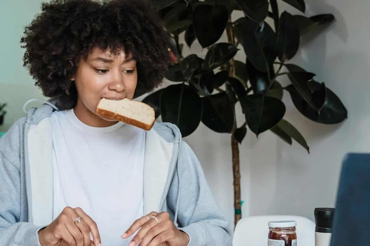 Woman with curly afro hair eating toast while wearing white shirt and gray hoodie, with houseplant in background