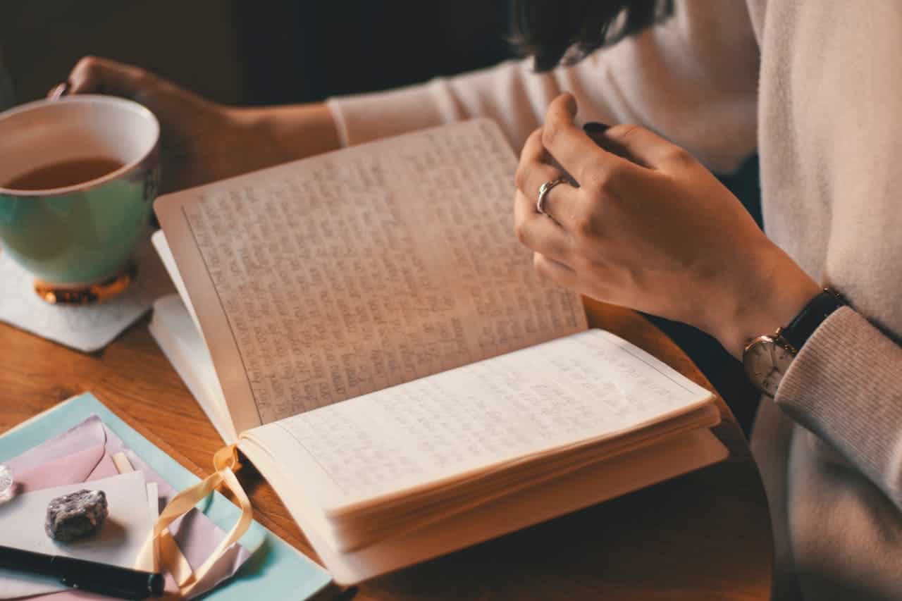 A person holding a cup of tea with one hand while the other hand flips through a handwritten journal, the journal has neatly written text, resting on a wooden desk with envelopes, a pen, and a stone, soft light highlighting the cozy setting, indicating a peaceful and reflective moment