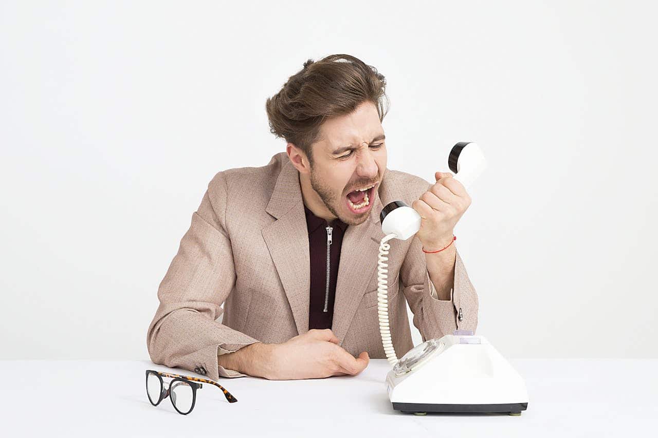 A man wearing a beige suit is shouting into an old-fashioned rotary phone, sitting at a desk with a pair of glasses nearby, showing frustration, his face expressing anger or irritation