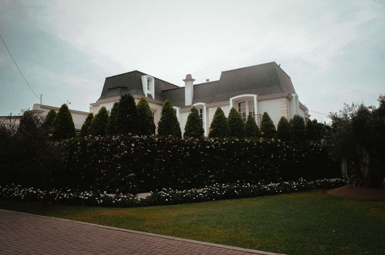 White mansion with dark mansard roof behind neatly trimmed cypress trees, flowering hedges, and lawn under overcast sky