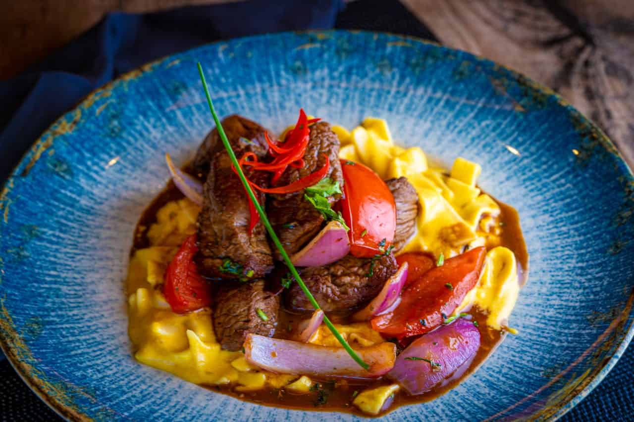A plate of tender beef served with sautéed vegetables, including tomatoes and onions, over a bed of pasta, garnished with fresh herbs and a red chili, arranged in a vibrant blue bowl with a glossy sauce