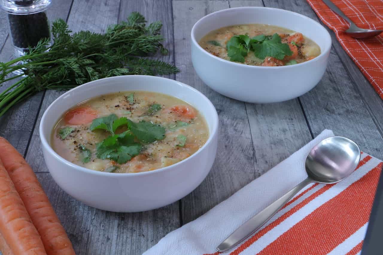 Two white bowls of chicken broth with vegetables and cilantro garnish, served with carrots and fresh herbs on wooden table