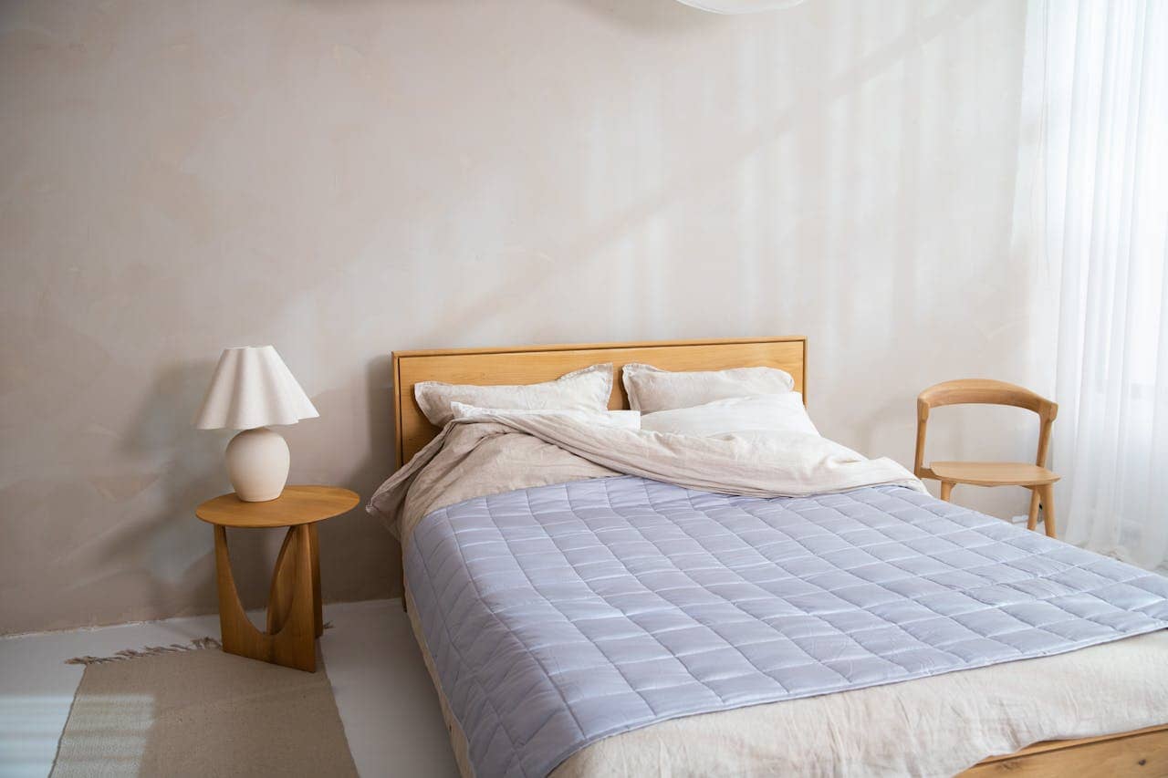 Minimalist bedroom with wooden bed frame, light blue mattress topper, and white pillows. Features wooden nightstand with ceramic lamp, wooden chair, white walls, and soft natural lighting from window