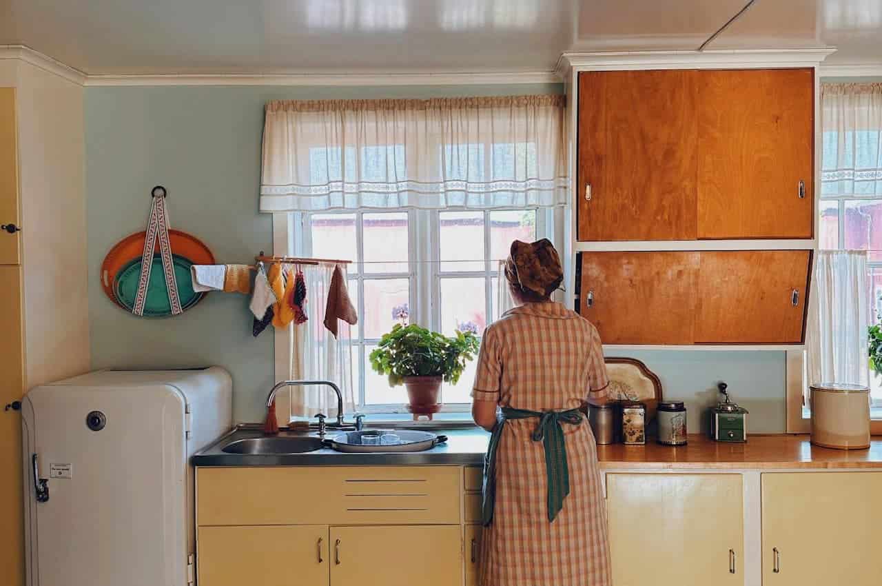 A person standing in a vintage kitchen, wearing a plaid dress and headscarf, washing dishes at the sink, dish towels hanging from a line next to the window, potted plant on the windowsill, retro yellow kitchen cabinets, and a white refrigerator to the left, warm and nostalgic atmosphere