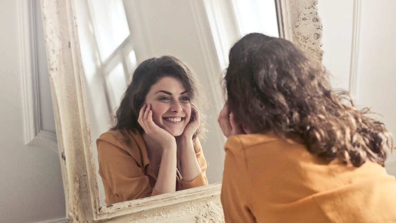 A woman sitting at a mirror, smiling, resting her chin on her hands, looking relaxed and content, in a cozy indoor setting, with a warm yellow top, reflecting on herself, enjoying her own company, calm expression, natural curly hair, peaceful ambiance, self-reflection moment