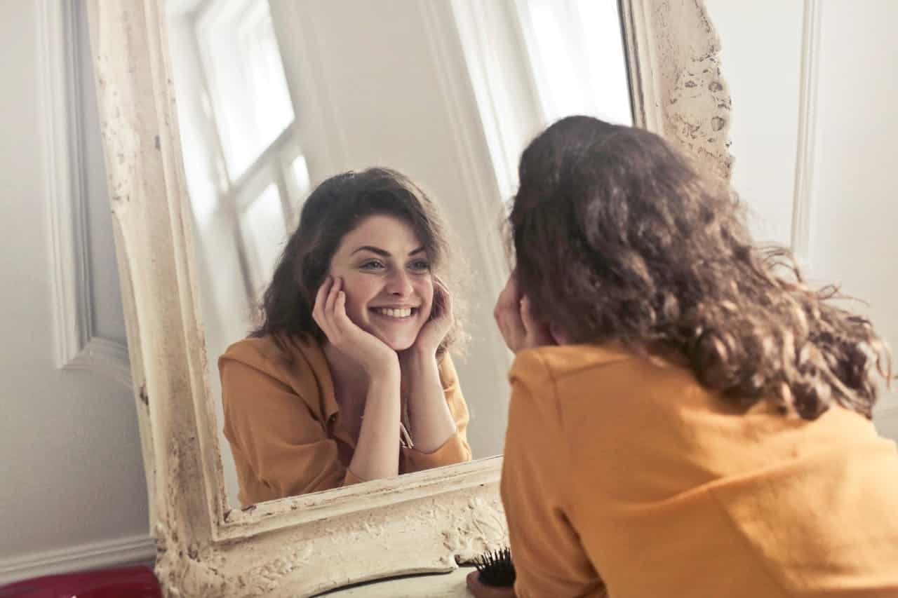 A woman sitting at a mirror, smiling, resting her chin on her hands, looking relaxed and content, in a cozy indoor setting, with a warm yellow top, reflecting on herself, enjoying her own company, calm expression, natural curly hair, peaceful ambiance, self-reflection moment