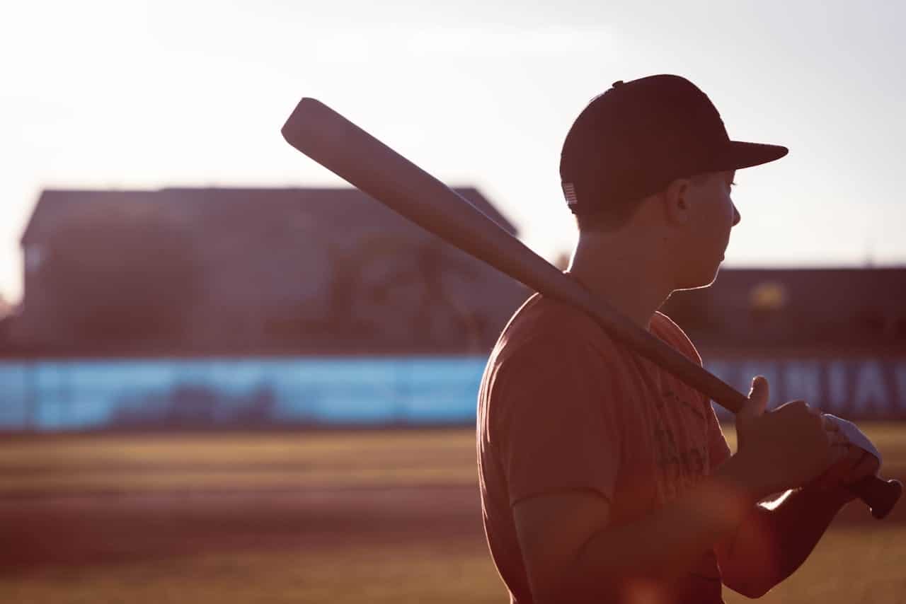 A young person holding a baseball bat over his shoulder, standing on a field, the sun is shining behind them, silhouette effect with a blurry background, the individual is wearing a cap and a t-shirt, giving a relaxed, ready stance in the setting of a baseball game