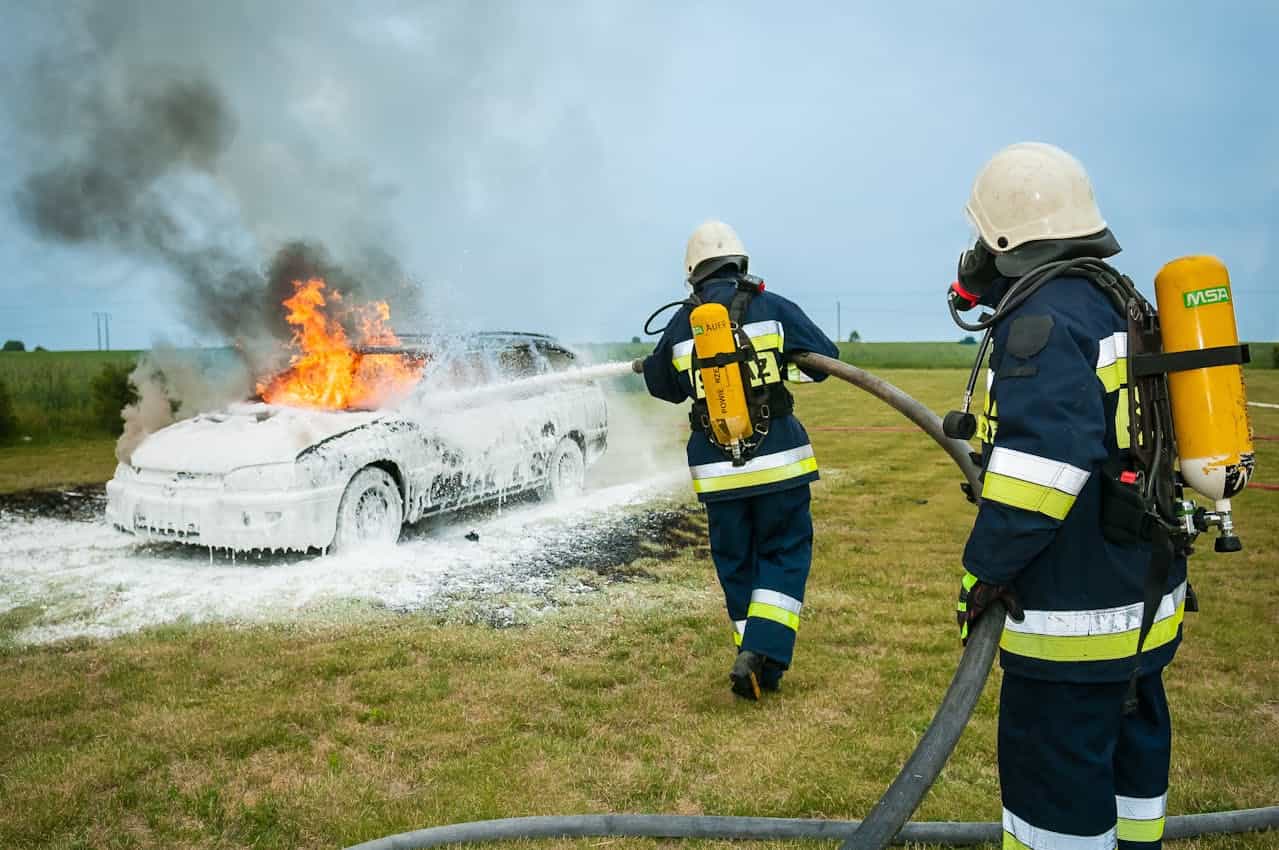 Two firefighters in protective gear are using a fire hose to spray foam on a burning car, thick smoke is rising from the fire, one firefighter is in the foreground, the other is behind, both are focused on extinguishing the flames, grassy field surrounding the scene, safety gear visible