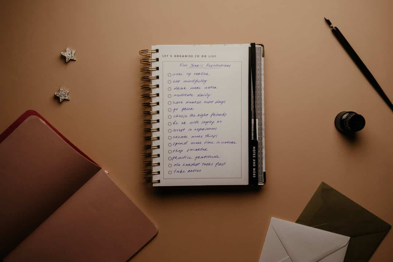 A flat lay image of a to-do list with things written in blue ink, resolutions include self-care goals like meditation, spending time in nature, and practicing gratitude, surrounded by a notebook, a pen, envelopes, and two star-shaped decorations