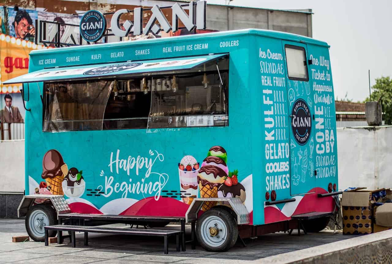 This is a colorful ice cream truck with the name "Giani," featuring a variety of desserts like ice cream, sundaes, kulfi, gelato, and shakes, the truck is brightly decorated with images of ice cream cones, with a cheerful "Happy Beginning" message, located in a bustling outdoor area with tables outside