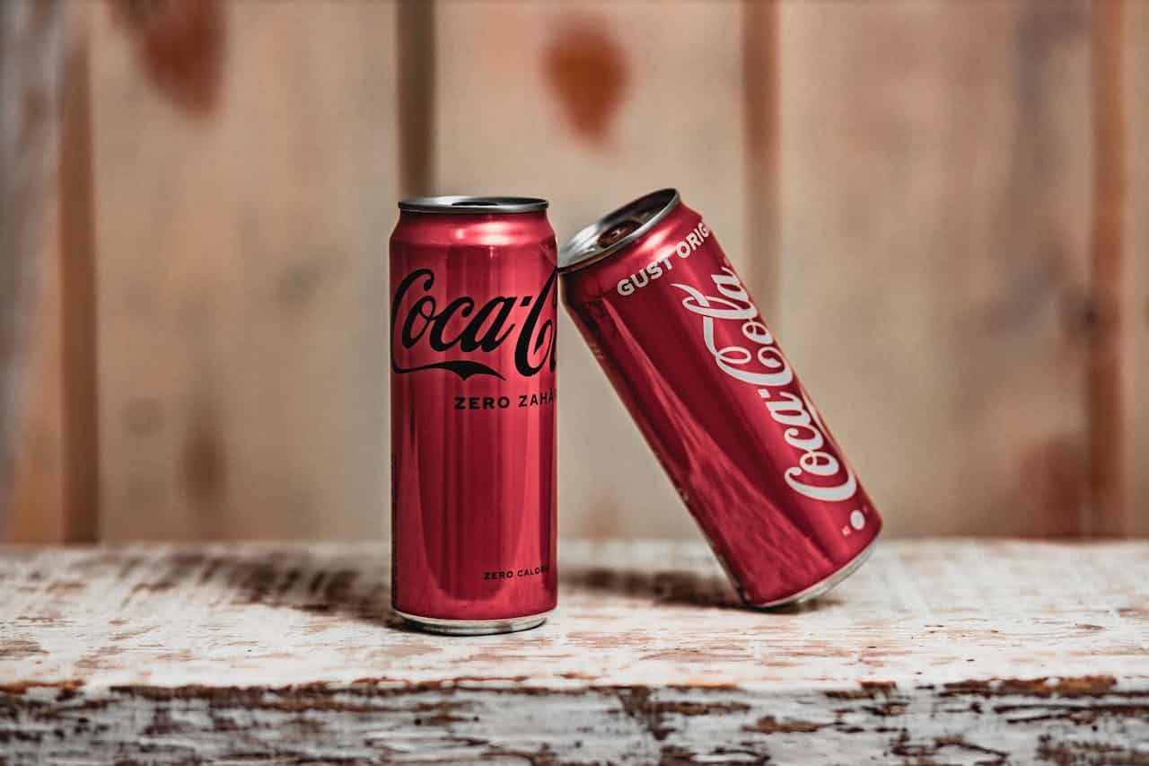 Alt text: Two Coca-Cola cans, one labeled "Coca-Cola Zero Zahár" and the other labeled "Coca-Cola Gust Original," leaning against each other on a rustic wooden surface, red cans with distinct branding visible, background softly blurred